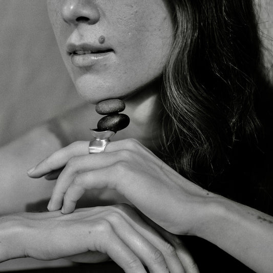 A close-up black and white photograph of a woman wearing a distinctive ring with stacked oval stones on her ring finger, with her hands resting near the frame.
