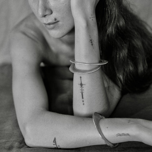 A black and white close-up of a woman resting with her arm bent, wearing several silver-toned bangles on her wrist and forearm, including a circular stack and a larger curved cuff.