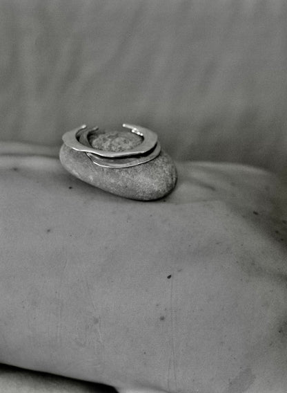 A silver ring with a textured circular top and a small stone inlay, displayed on a smooth gray stone against a soft gray fabric background.