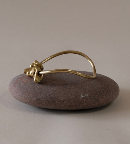 A polished brass bangle with charm pebbles. displayed on a smooth, round brown stone against a neutral beige background.