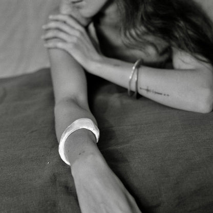 A black and white close-up of a person lying down with one arm raised, wearing a wide, chunky silver cuff bracelet on the wrist.